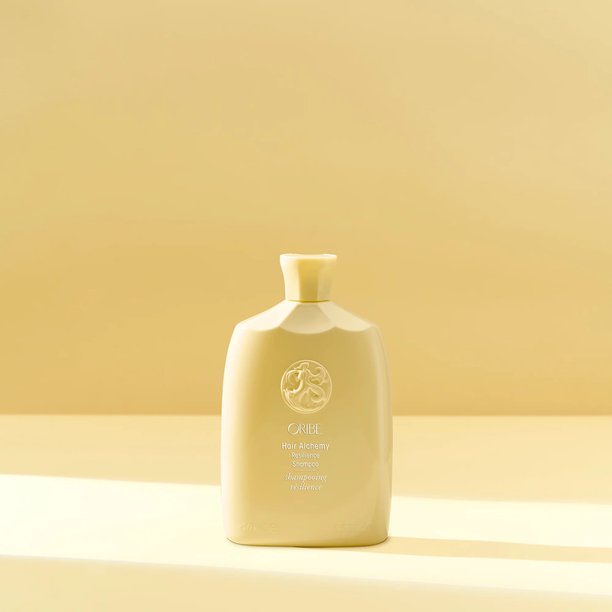 A pale yellow bottle of Oribe Hair Alchemy Resilience Shampoo with a floral design near the cap.