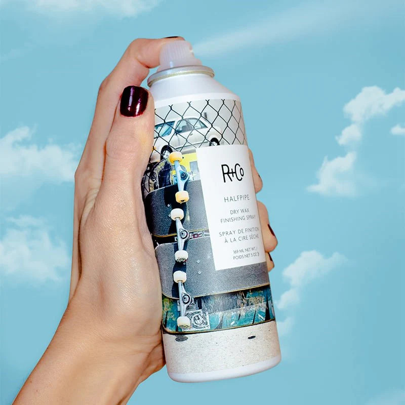 A hand holding a can of R+Co Halfpipe Dry Wax Finishing Spray with a blue sky and clouds in the background.