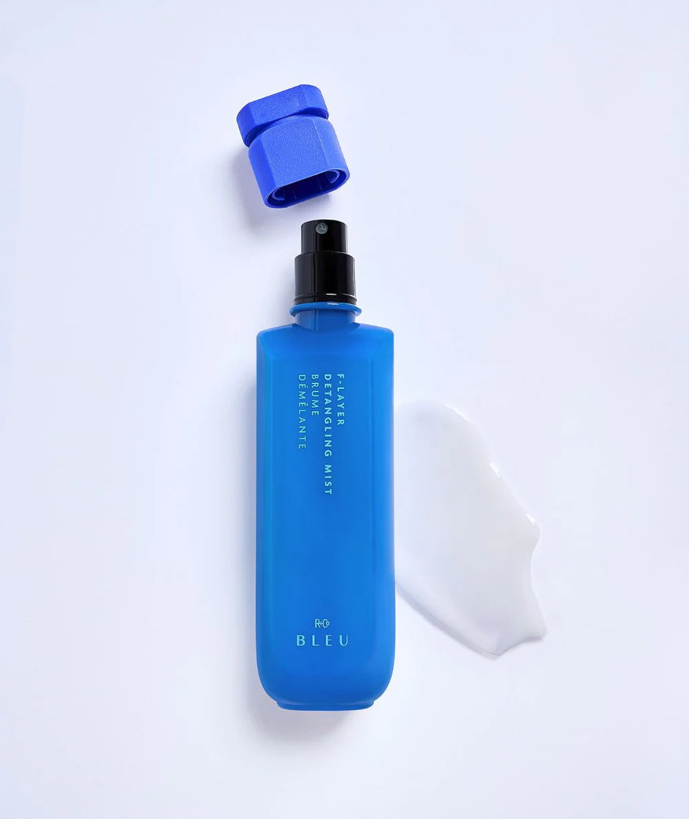 A blue bottle of R+Co F-Layer Detangling Mist with its cap removed, next to a white cloth, on a white background.