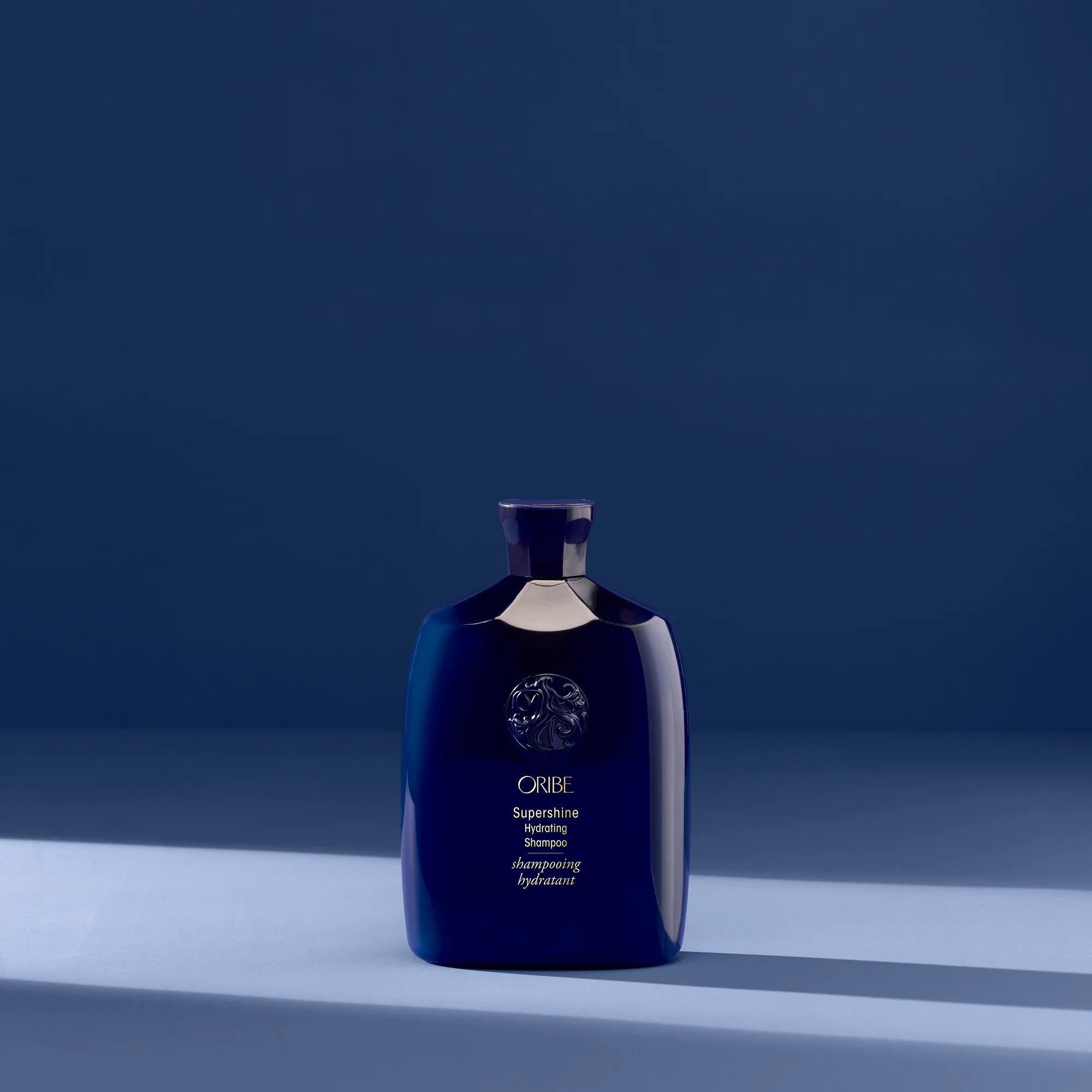 A bottle of Oribe Supershine Shampoo with a dark blue label on a blue background.