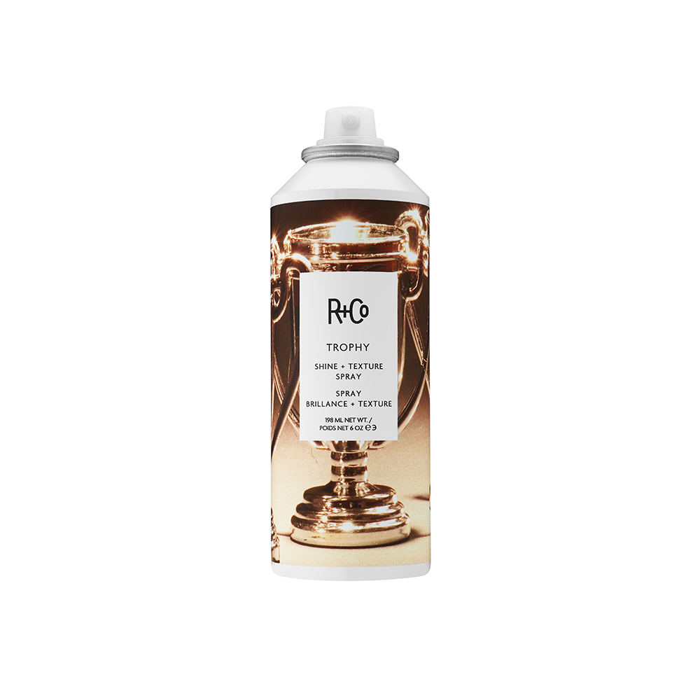 A bottle of R+Co Trophy Shine & Texture Spray with a white and gold design, indicating it is a hair styling product.