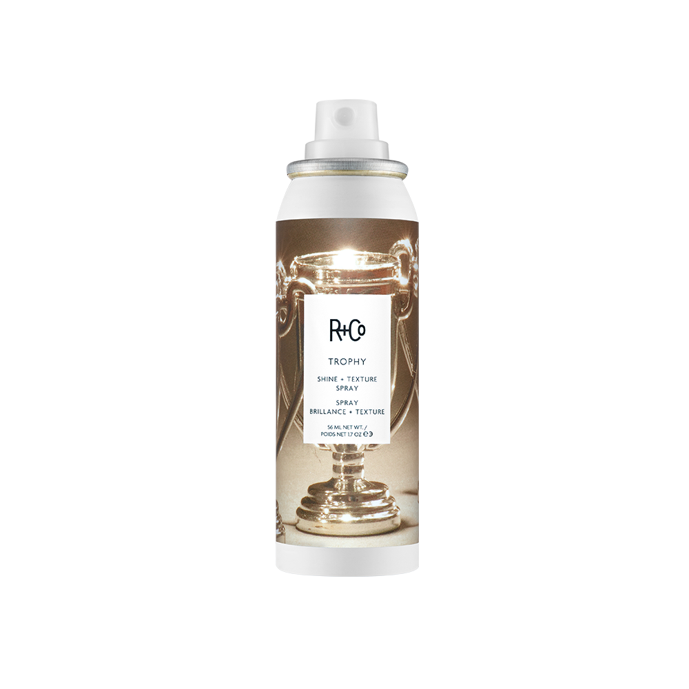 A bottle of R+Co Trophy Shine & Texture Spray with a white and gold design, indicating it is a hair styling product.