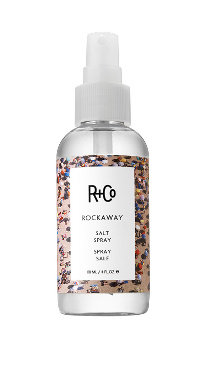A spray bottle of R+Co Rockaway Salt Spray with a white label and decorative patterns.